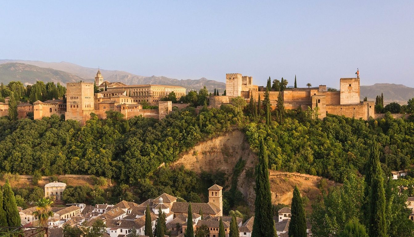 Alhambra fortress and palaces atop a wooded hill at golden hour, with Albaicín rooftops and cypresses below