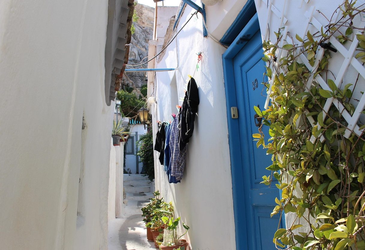 Narrow whitewashed alley with a blue door, potted plants and hanging laundry beneath the Acropolis rock