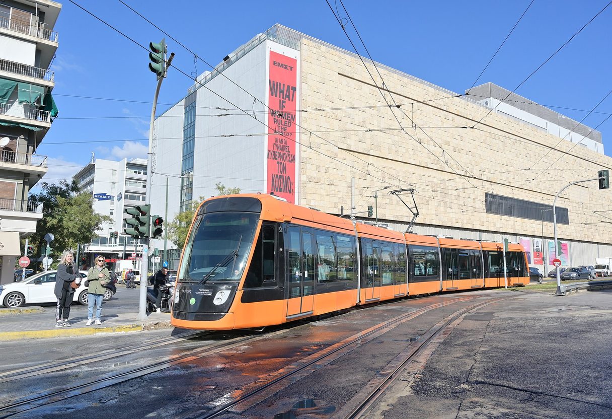 Orange Alstom Citadis tram curving along tracks on a sunny Athens street with pedestrians nearby