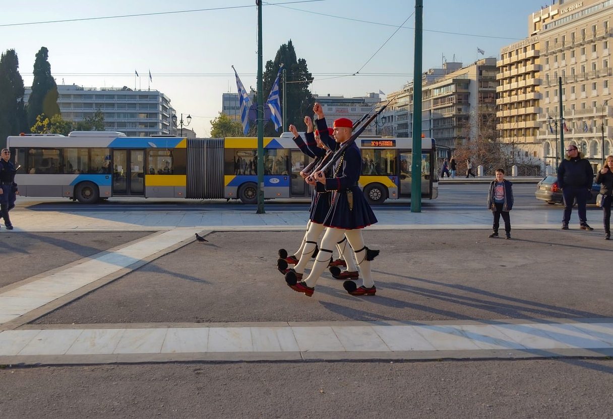 Evzone guards marching near Syntagma Square as a yellow and blue Athens trolleybus passes behind