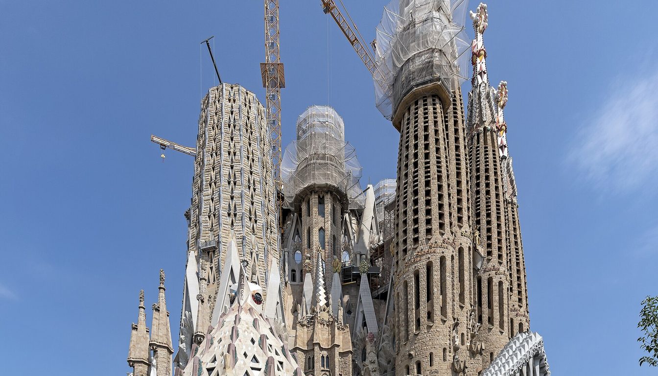 Exterior towers of the Sagrada Família apse under blue sky, with construction cranes and street in front
