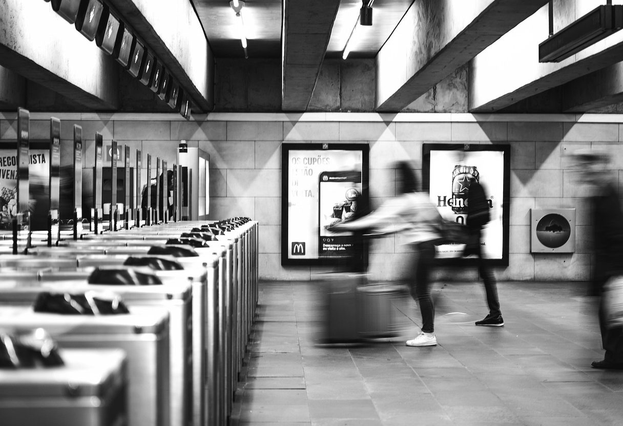 Black and white metro station with a row of ticket gates and blurred commuters pulling luggage past