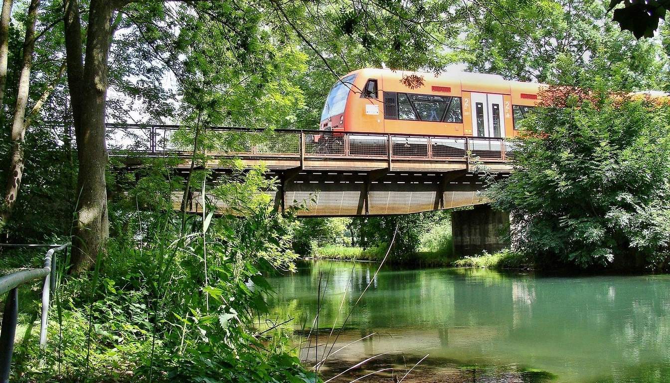 Orange regional train crossing a small railway bridge over a clear green river framed by lush trees