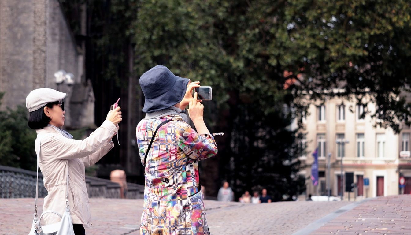 Two tourists holding up smartphones to take photos on a cobbled square lined with historic European buildings