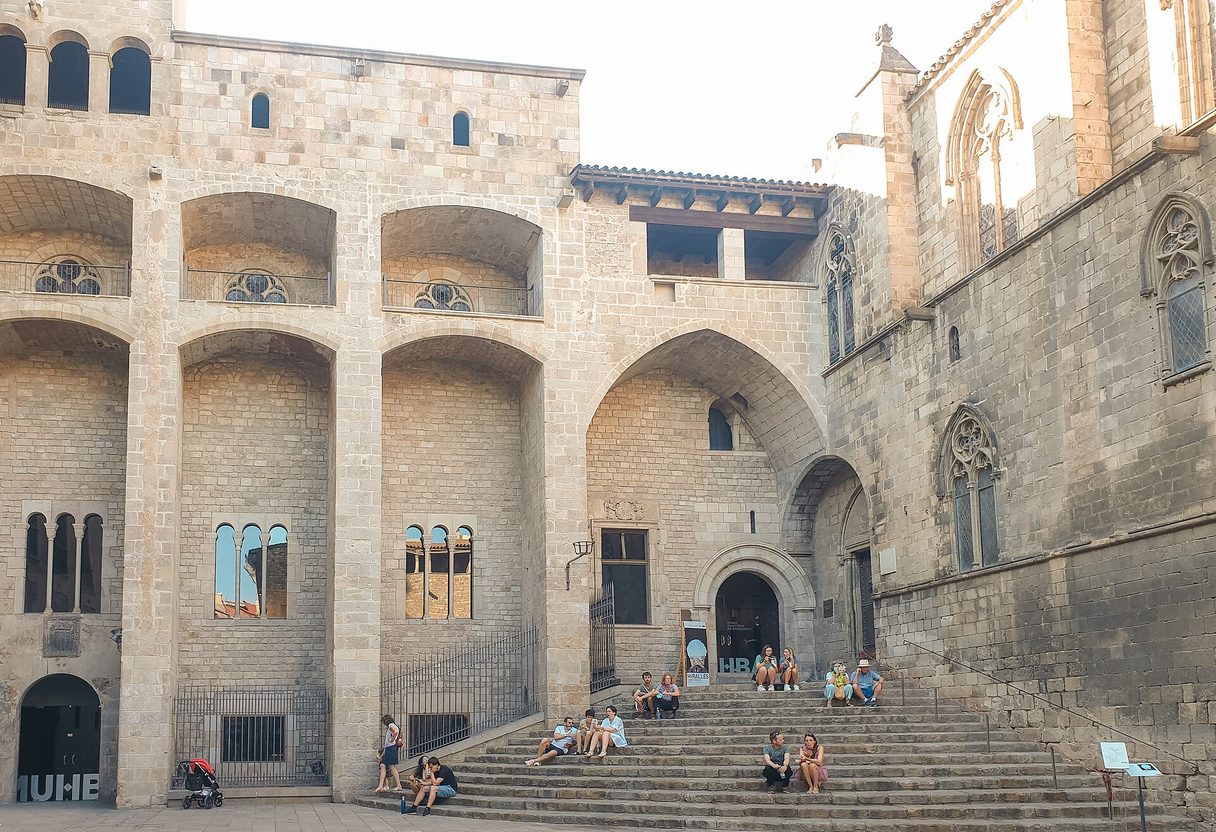 Stone steps and arched facade of the Palau Reial Major in Plaça del Rei, with people sitting on the stairs