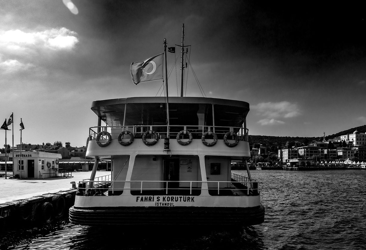 Black-and-white view of an Istanbul ferry moored at the island pier with waterfront buildings behind