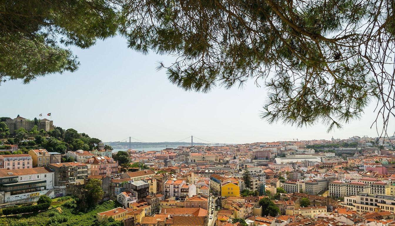 Red rooftops of Lisbon framed by pine branches, with São Jorge Castle and the 25 de Abril Bridge over the Tagus beyond