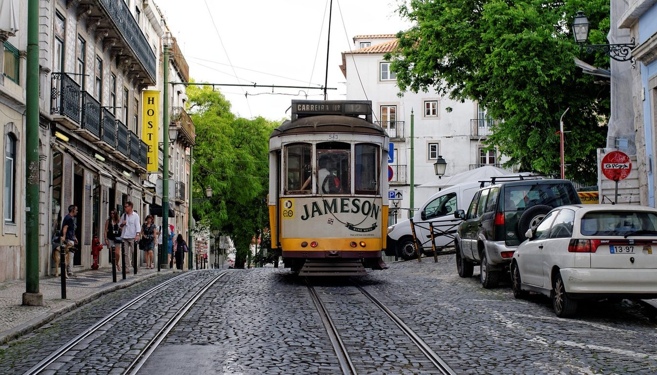 Classic yellow Lisbon tram descending a sloping cobbled street lined with old buildings, trees and parked cars