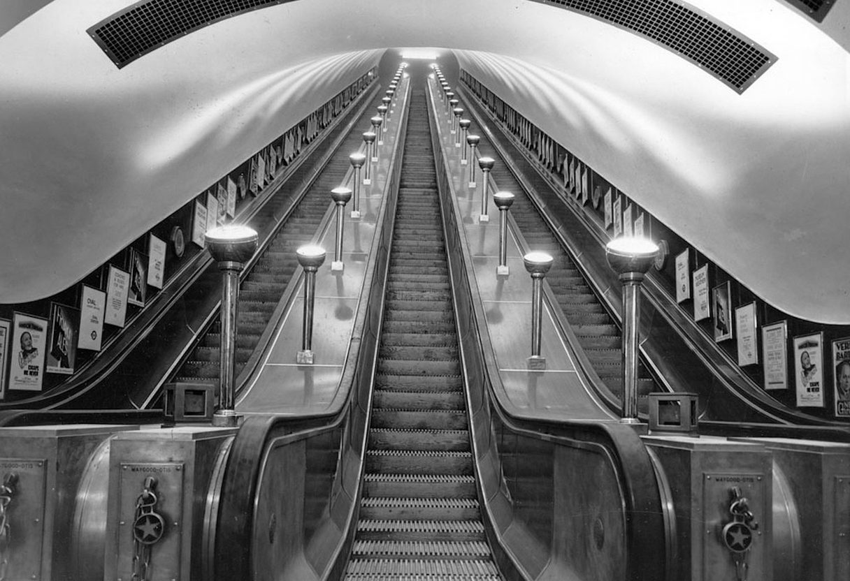 Black-and-white view up a long Tube escalator shaft with uplighter lamps and posters lining the curved tunnel walls
