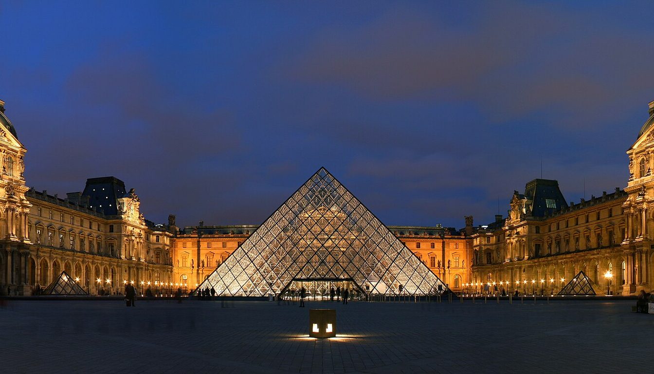 Illuminated Louvre pyramid at night in front of the golden-lit palace facade under a deep blue sky