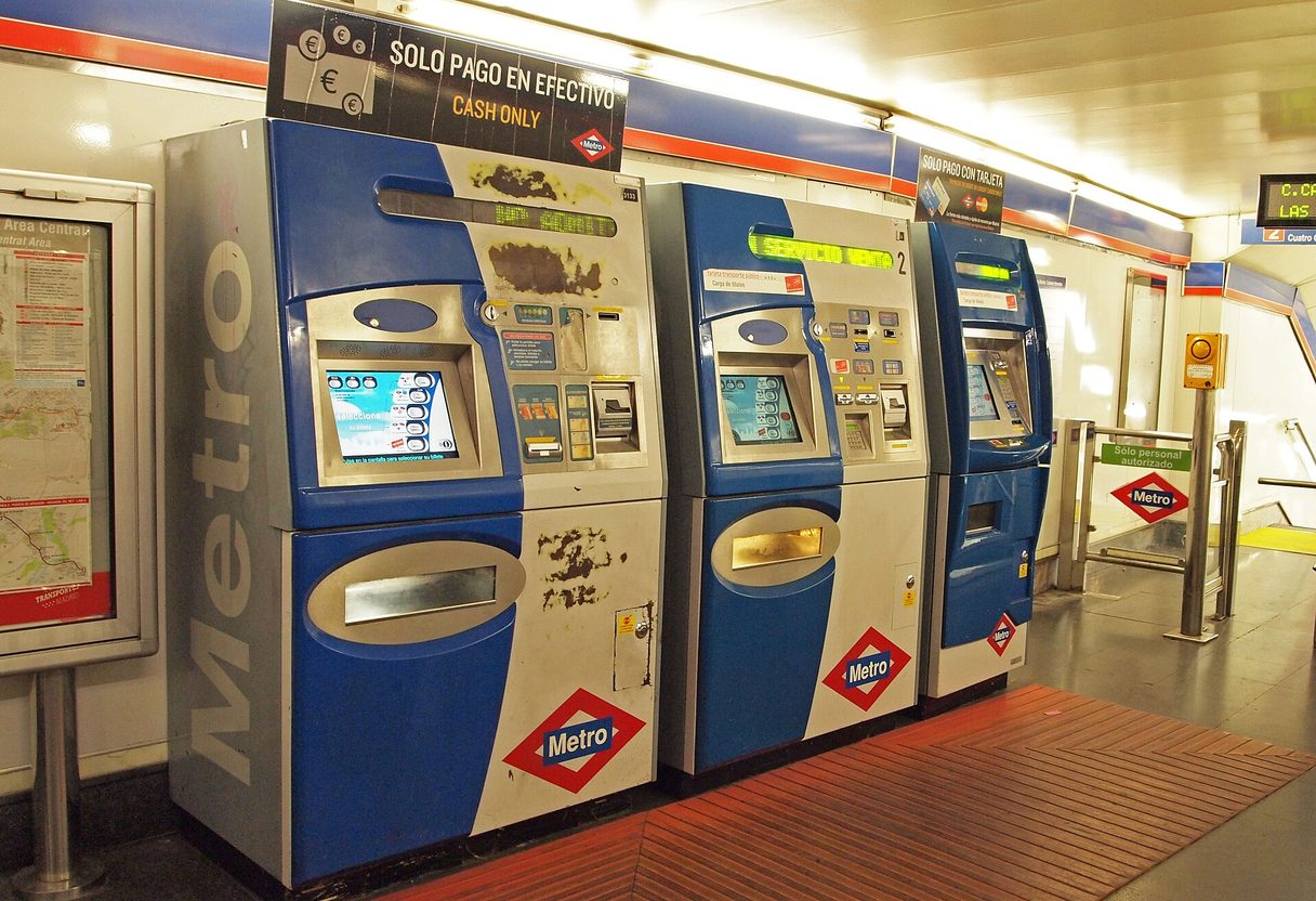 Row of blue and white Metro de Madrid ticket machines in a station corridor, one marked cash only, next to ticket gates