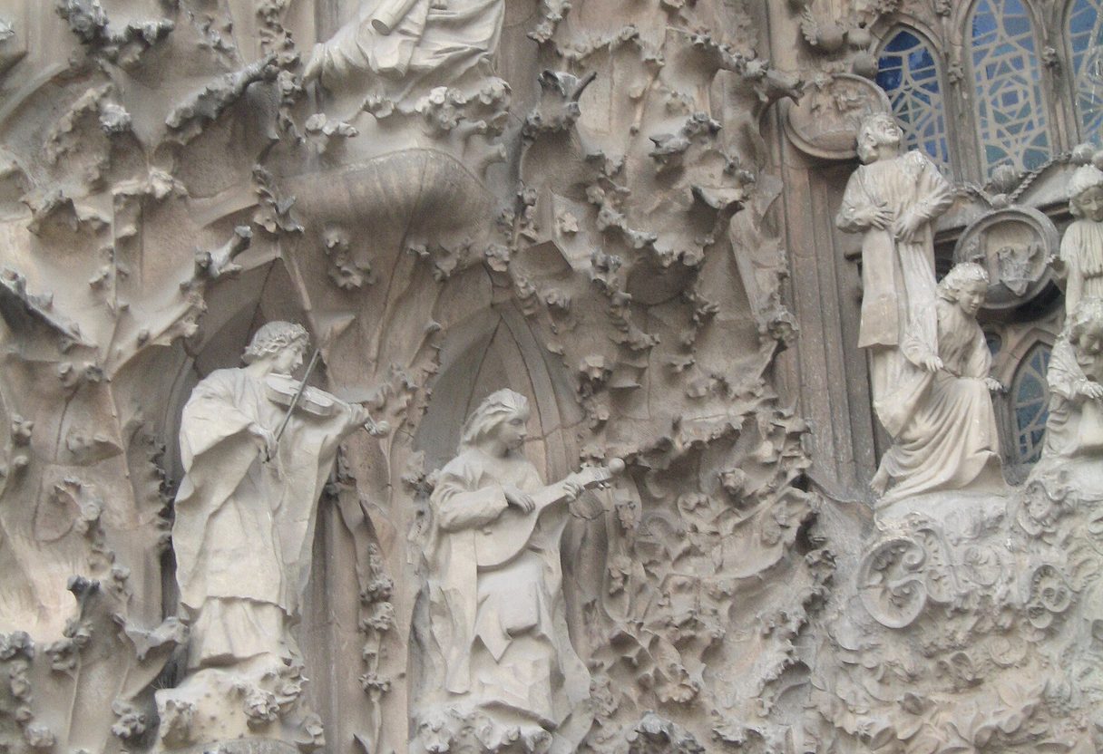 Close-up of the Nativity façade with sculpted angels playing instruments amid dense stone foliage and a stained-glass window