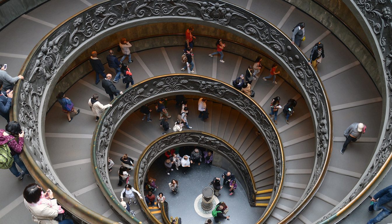 Crowds of visitors walking down the Vatican Museums spiral staircase, seen from above with its decorative railing