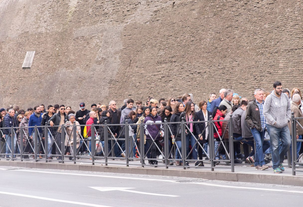 Crowd of visitors queuing behind metal railings along the brick Vatican wall outside the Vatican Museums