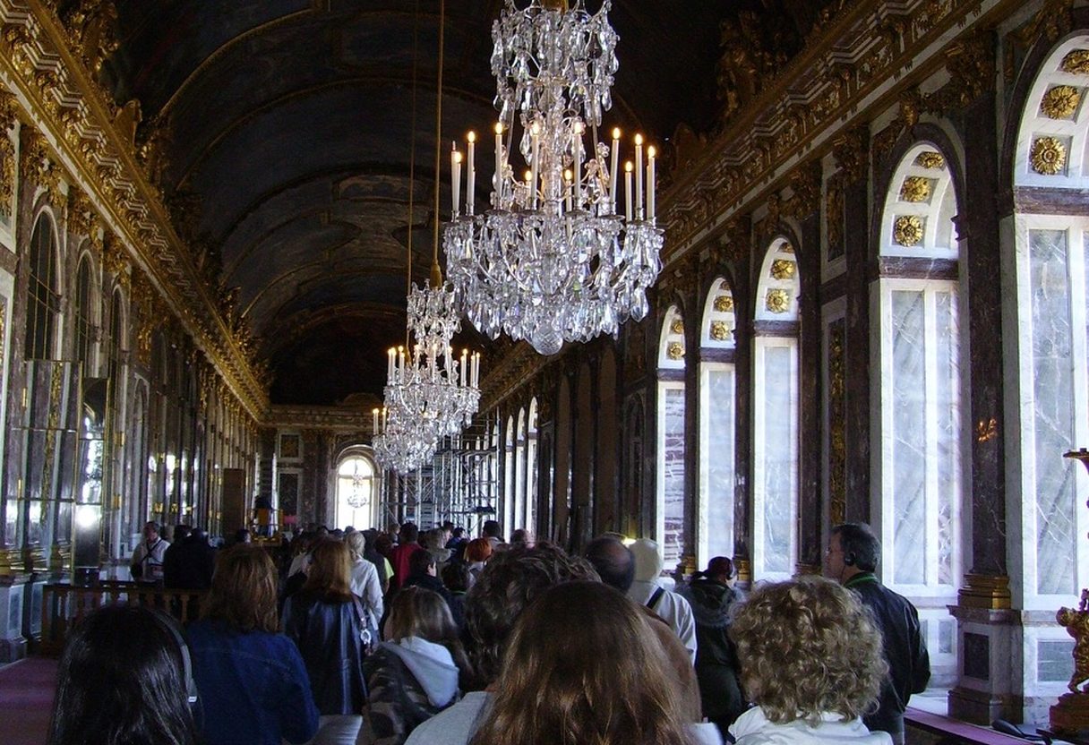 Crowds of tourists filling the Hall of Mirrors at Versailles beneath crystal chandeliers and a painted vaulted ceiling
